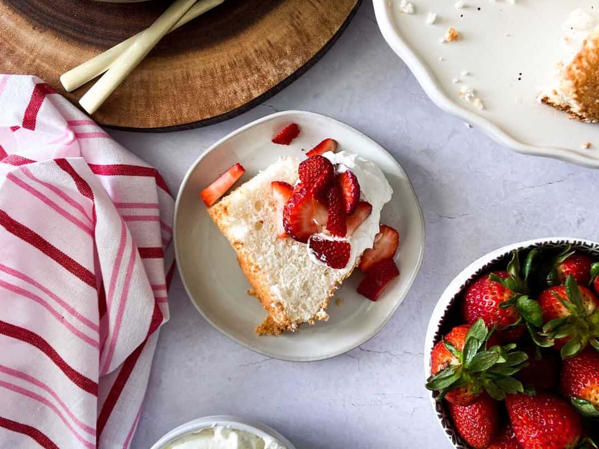 angel food cake topped with whipped cream and strawberries next to rest of cake, bowl of whipped cream, and bowl of strawberries