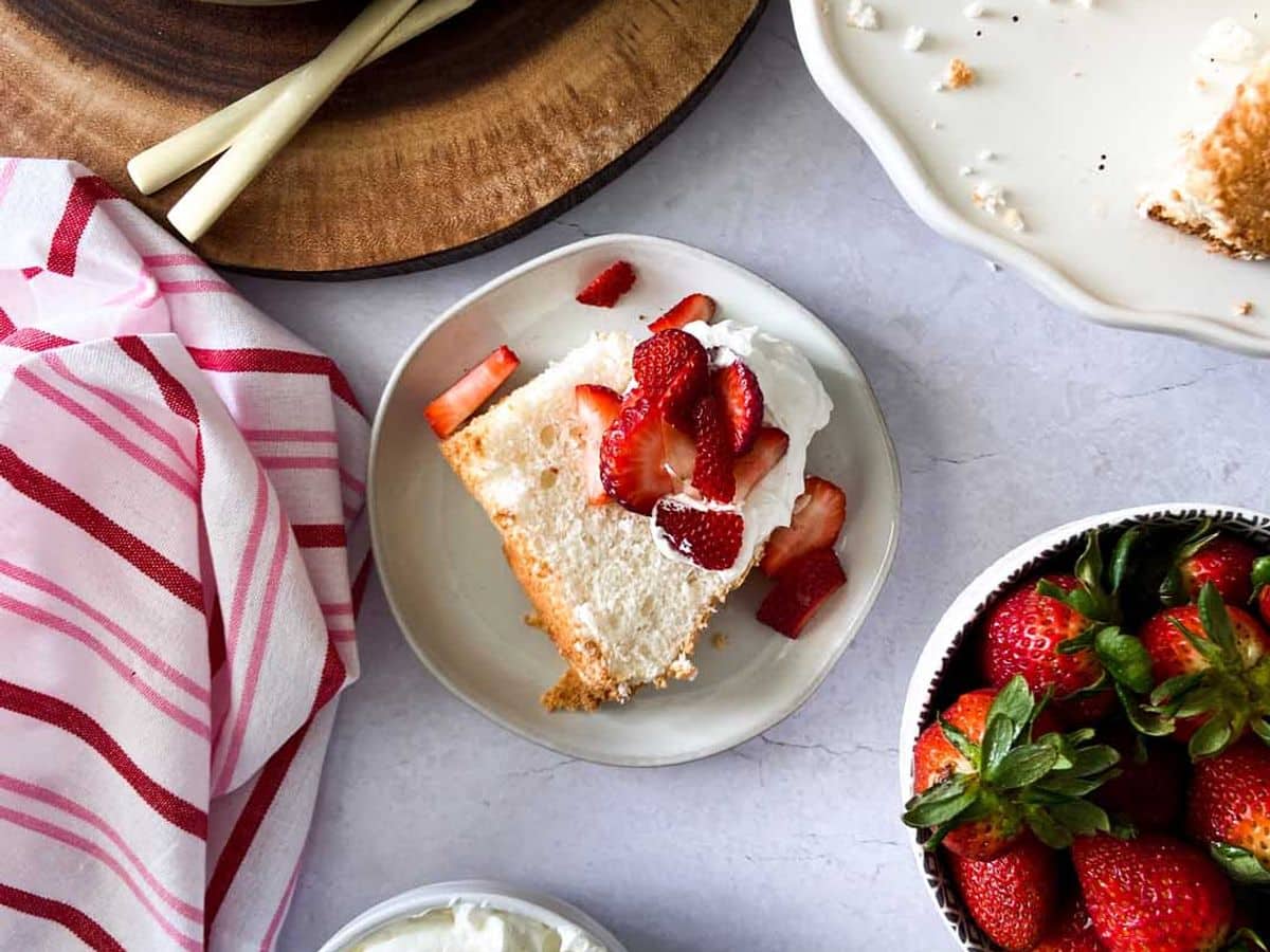 angel food cake topped with whipped cream and strawberries next to rest of cake, bowl of whipped cream, and bowl of strawberries
