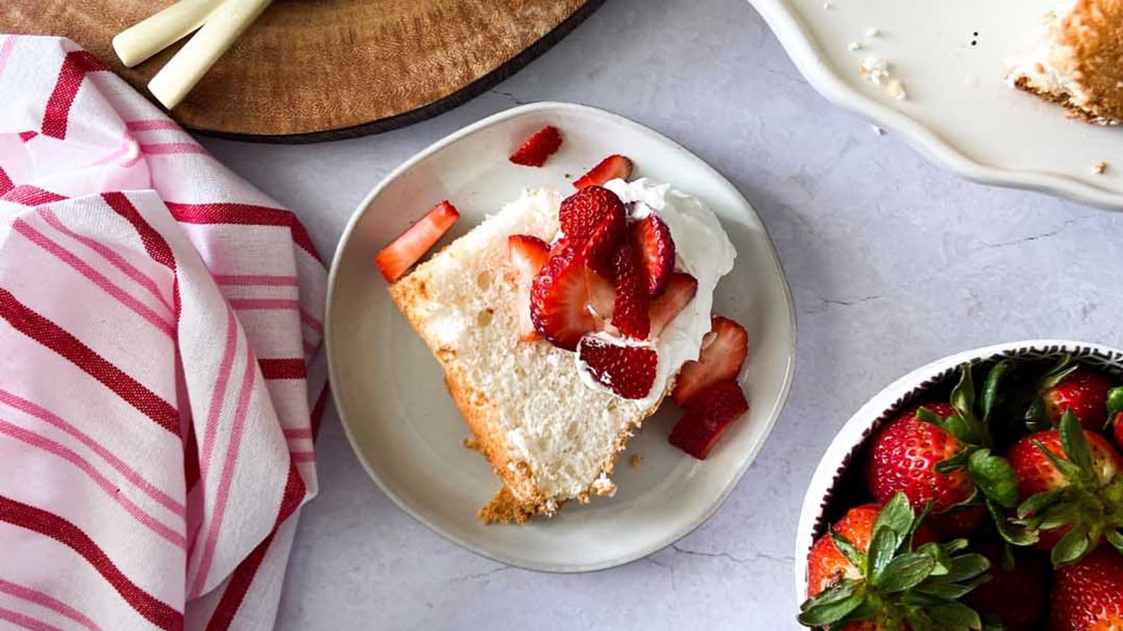 angel food cake topped with whipped cream and strawberries next to rest of cake, bowl of whipped cream, and bowl of strawberries