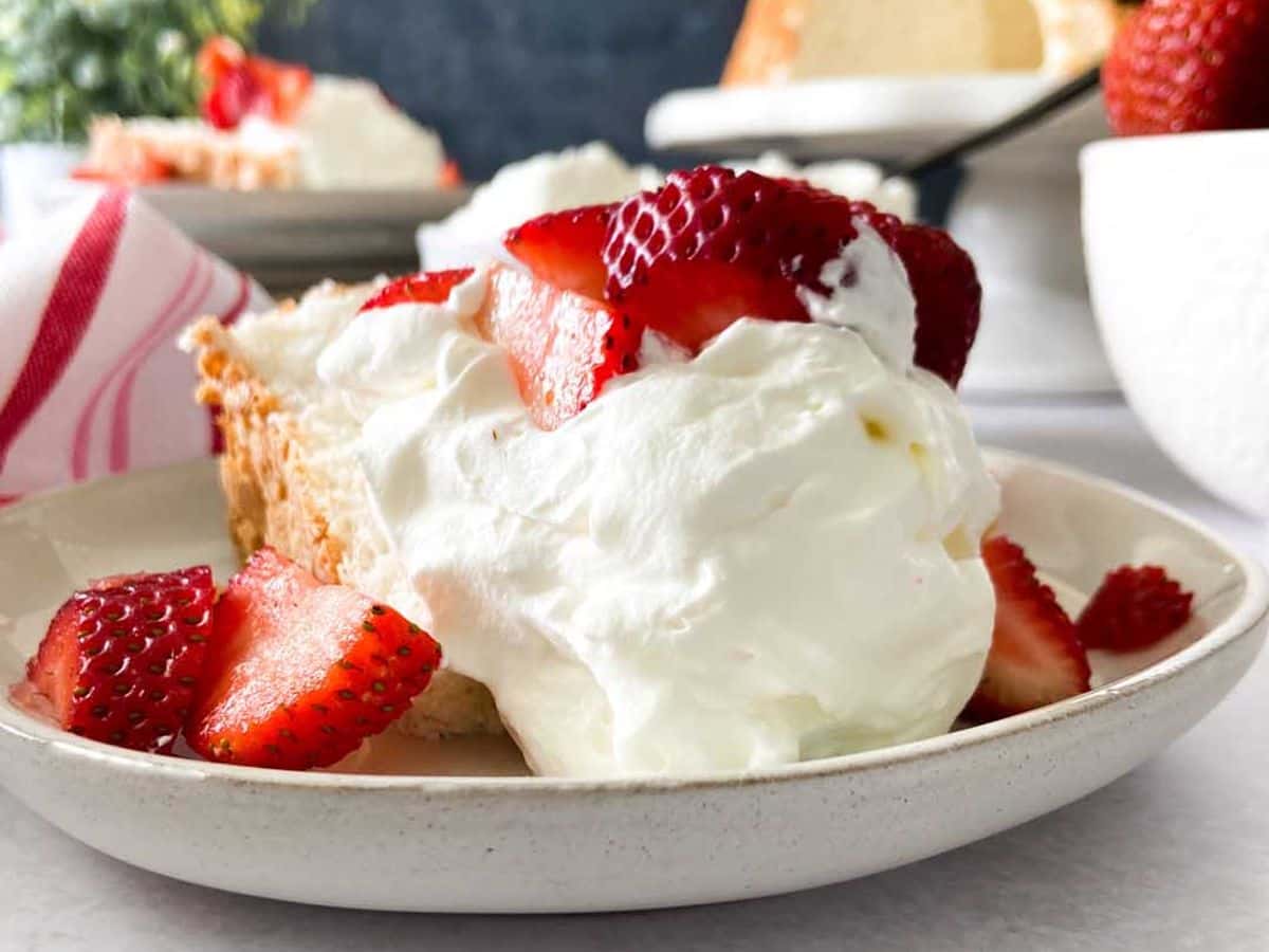 angel food cake on a plate with whipped cream and strawberries with the rest of the cake in the background
