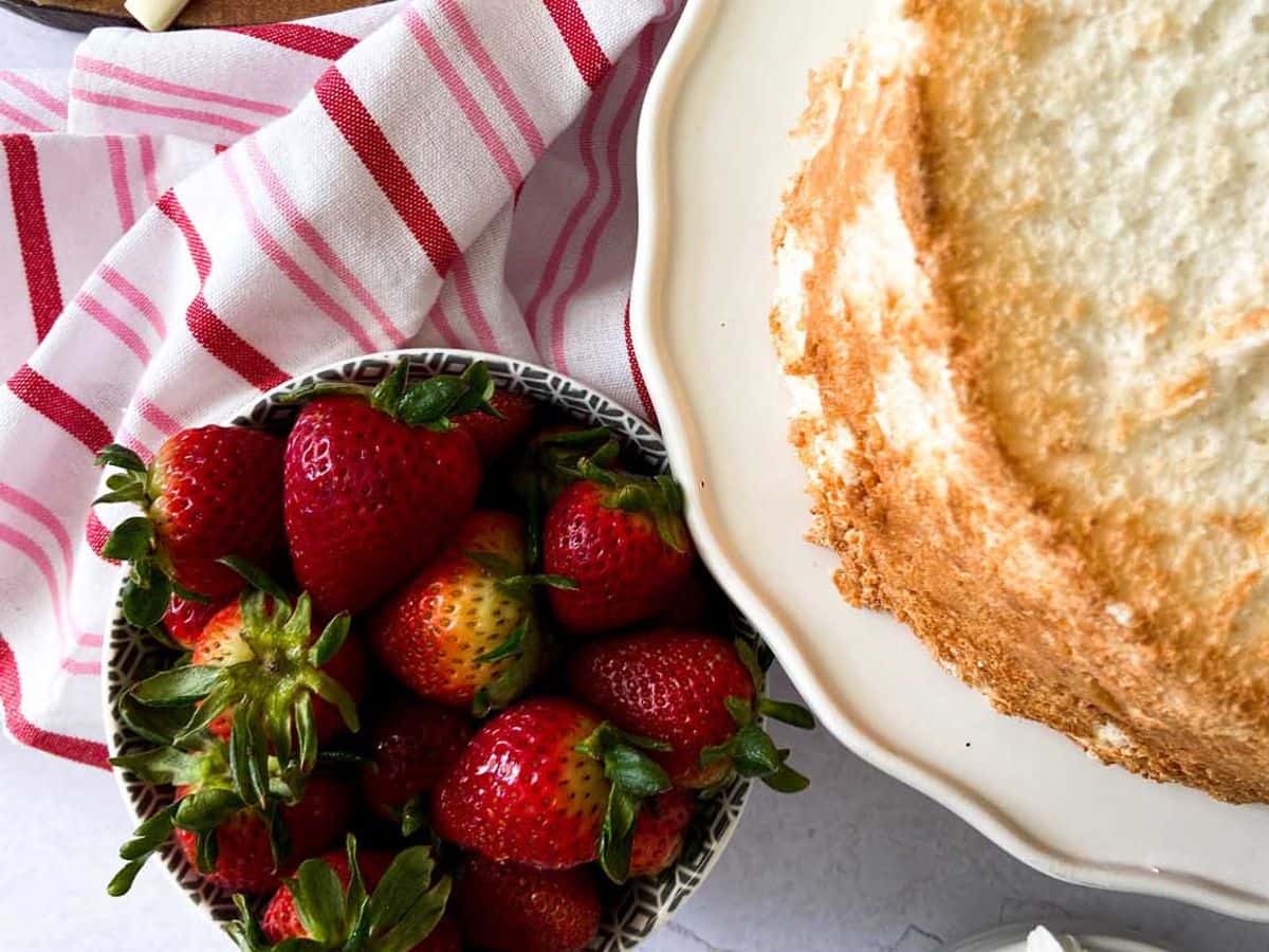 angel food cake whole cake finished next to a bowl of whipped cream and a bowl of strawberries