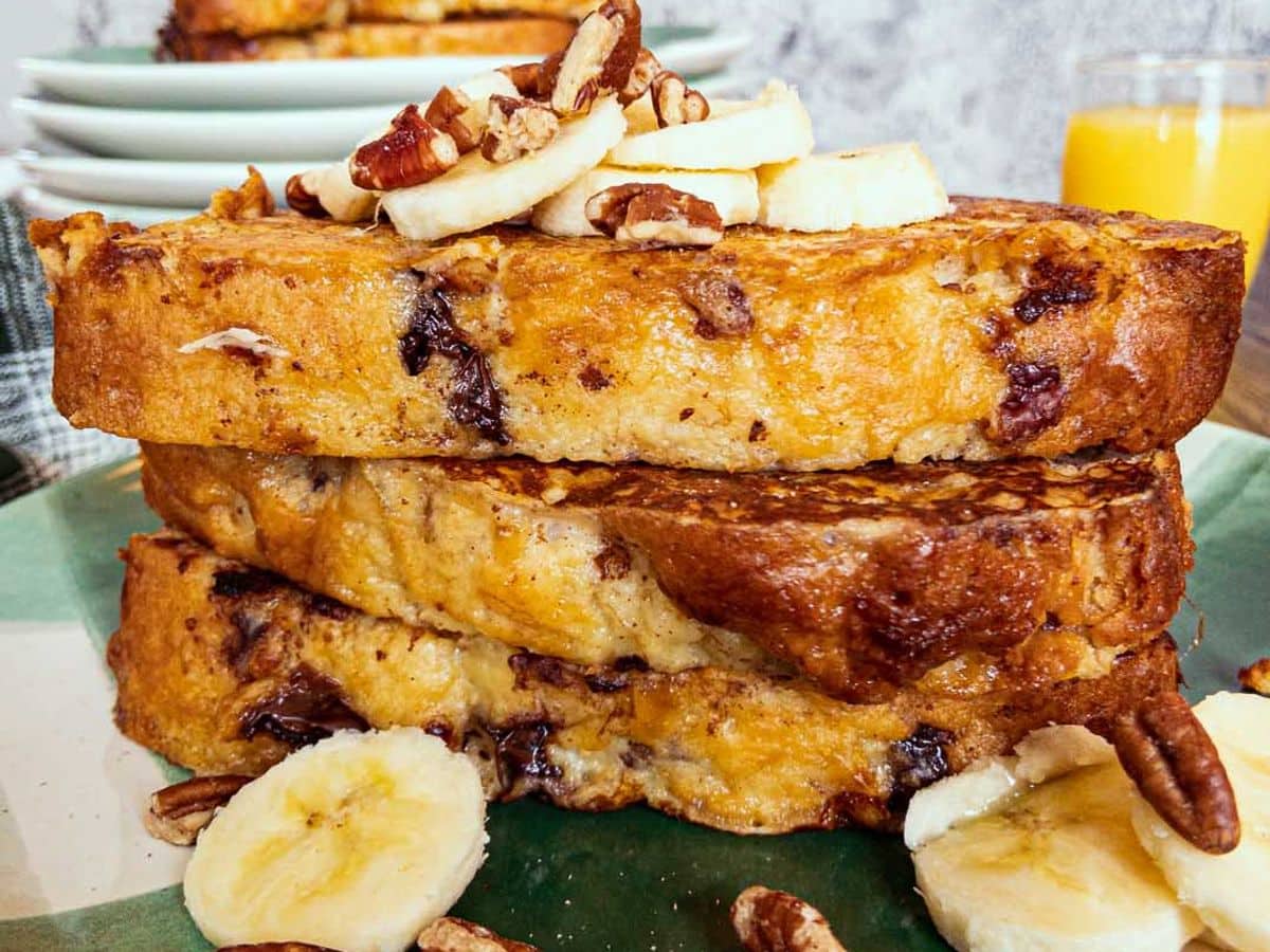 banana bread french toast stacked and topped with sliced banana, and nuts, with a glass of orange juice in the background