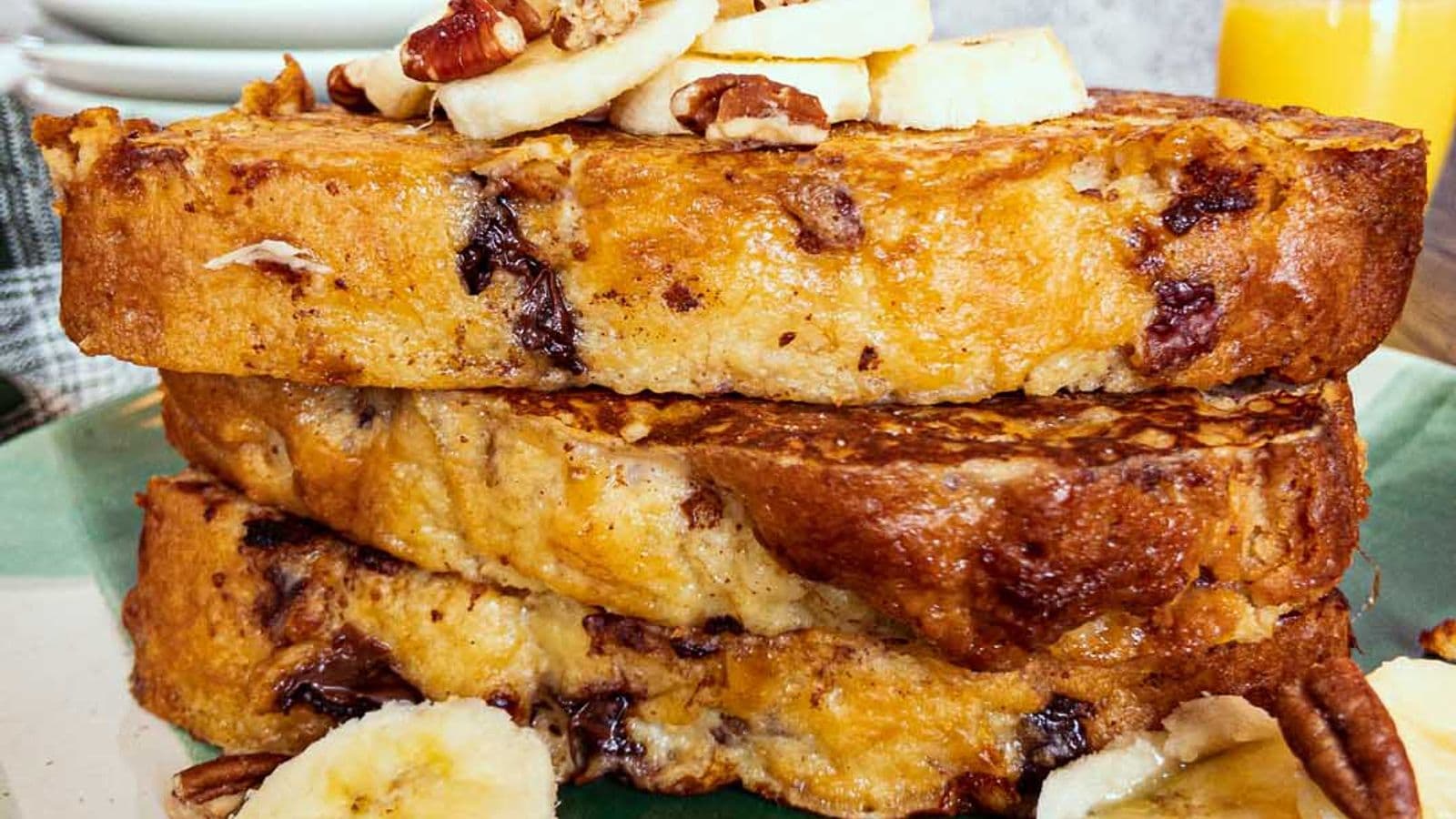 banana bread french toast stacked and topped with sliced banana, and nuts, with a glass of orange juice in the background