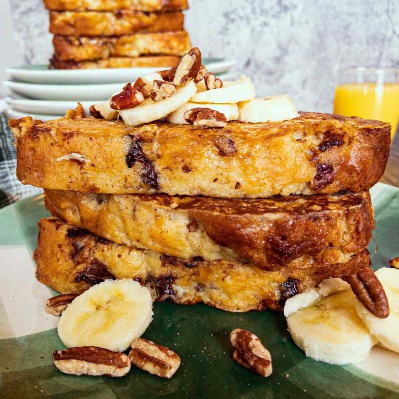 banana bread french toast stacked and topped with sliced banana, and nuts, with a glass of orange juice in the background