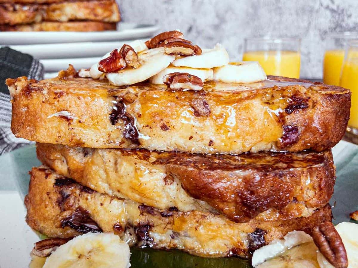banana bread french toast stacked and topped with sliced banana, nuts, and syrup, with glasses of orange juice in the background