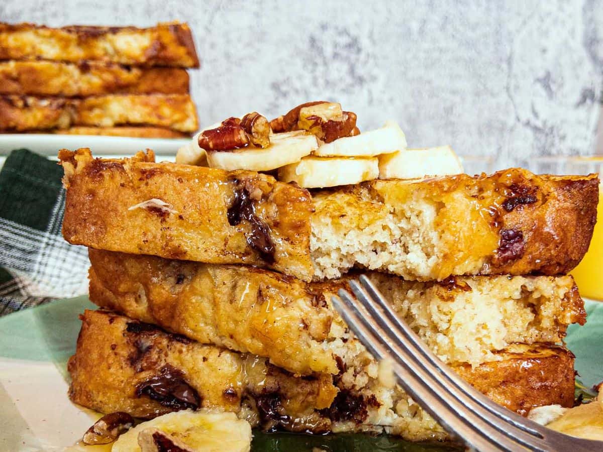 banana bread french toast stacked and topped with sliced banana, nuts, and syrup, with bite taken out of the front and a fork