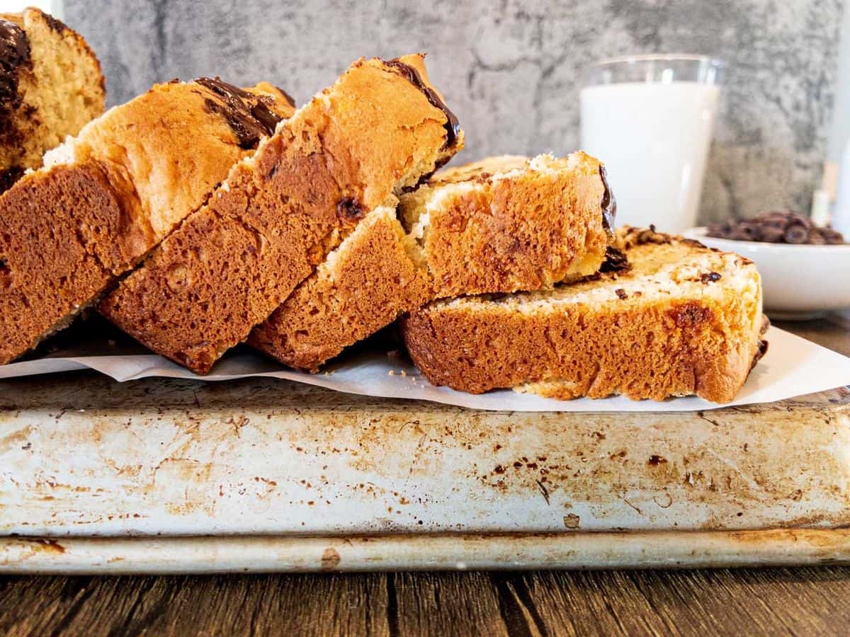 banana bread with chocolate chips sliced with slices falling over and bowl of chocolate chips and glass of milk in the background
