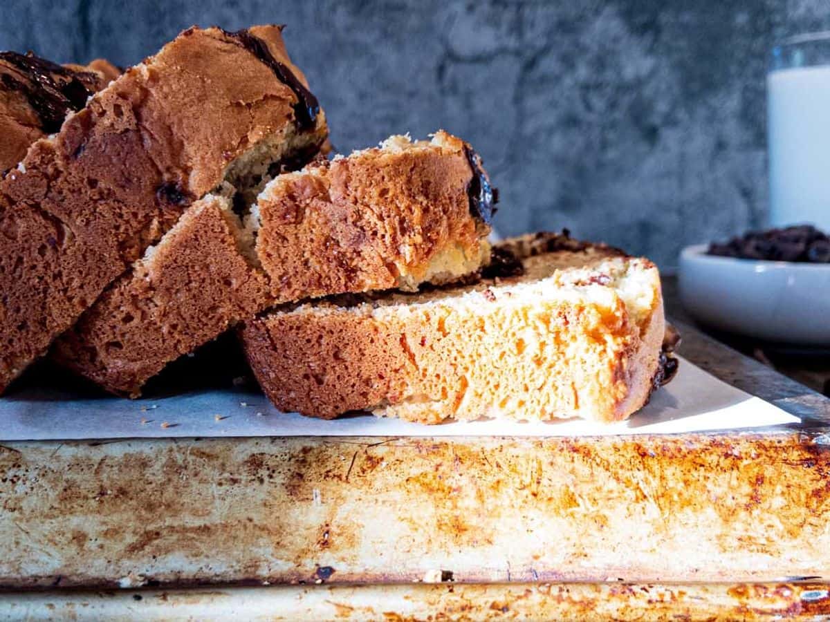 banana bread with chocolate chips, sliced on the back of a cookie sheet, with the slices falling over and chocolate chips and a glass of milk in the background