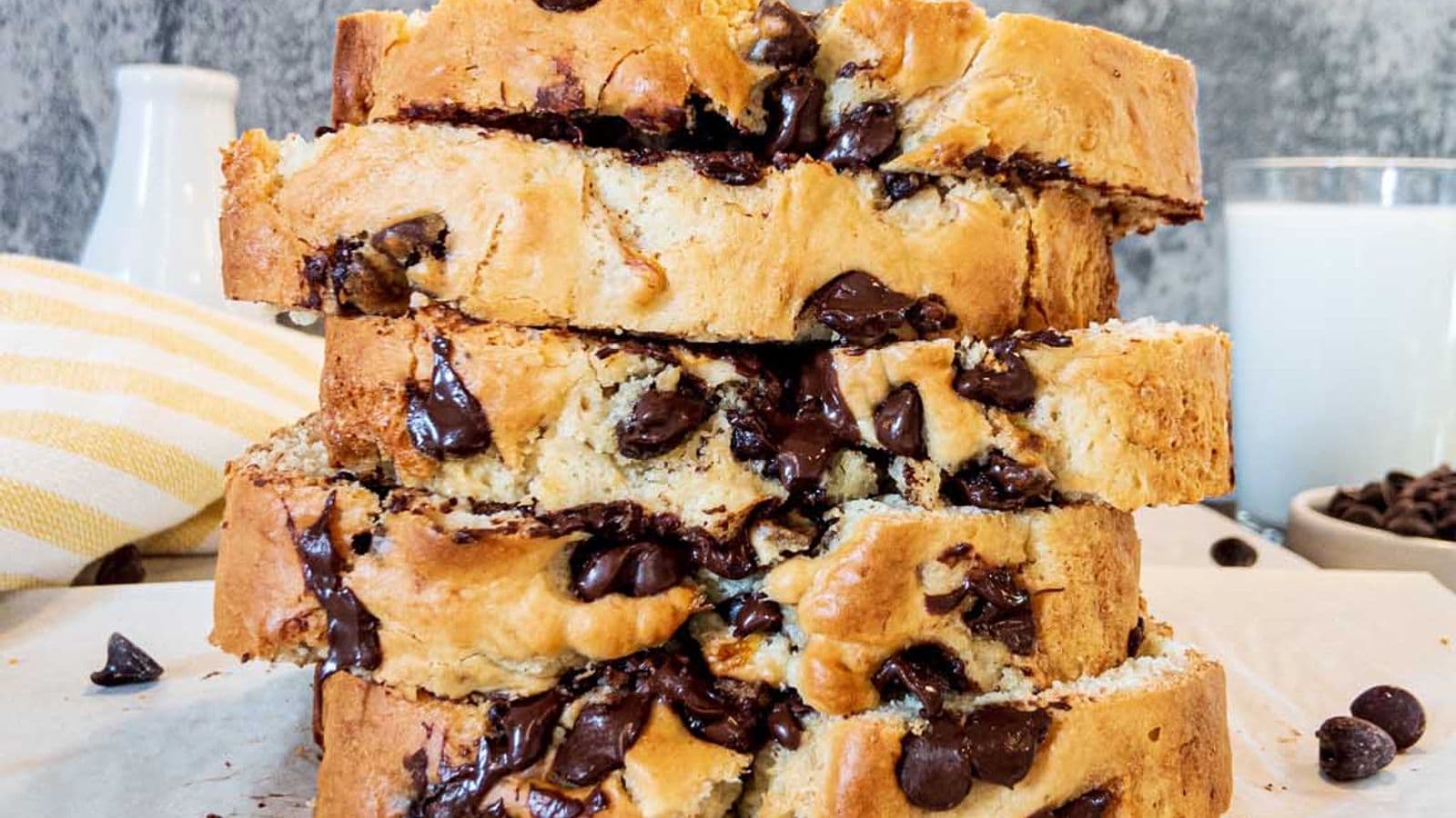 banana bread with chocolate chips, sliced and stacked on the back of a cookie sheet, with glass of milk and bowl of chocolate chips in the background
