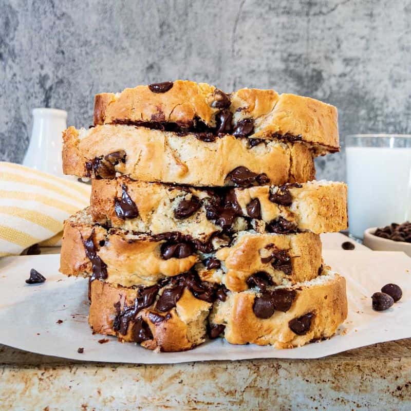 banana bread with chocolate chips, sliced and stacked on the back of a cookie sheet, with glass of milk and bowl of chocolate chips in the background