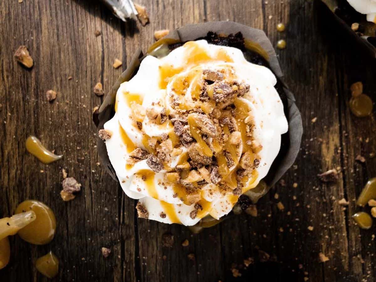 better than sex cupcakes topped with whipped cream, caramel sauce, and Heath bits, on the counter next to a piping bag filled with whipped cream