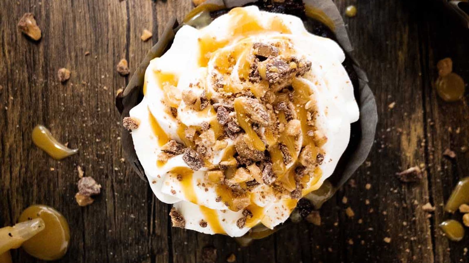 better than sex cupcakes topped with whipped cream, caramel sauce, and Heath bits, on the counter next to a piping bag filled with whipped cream