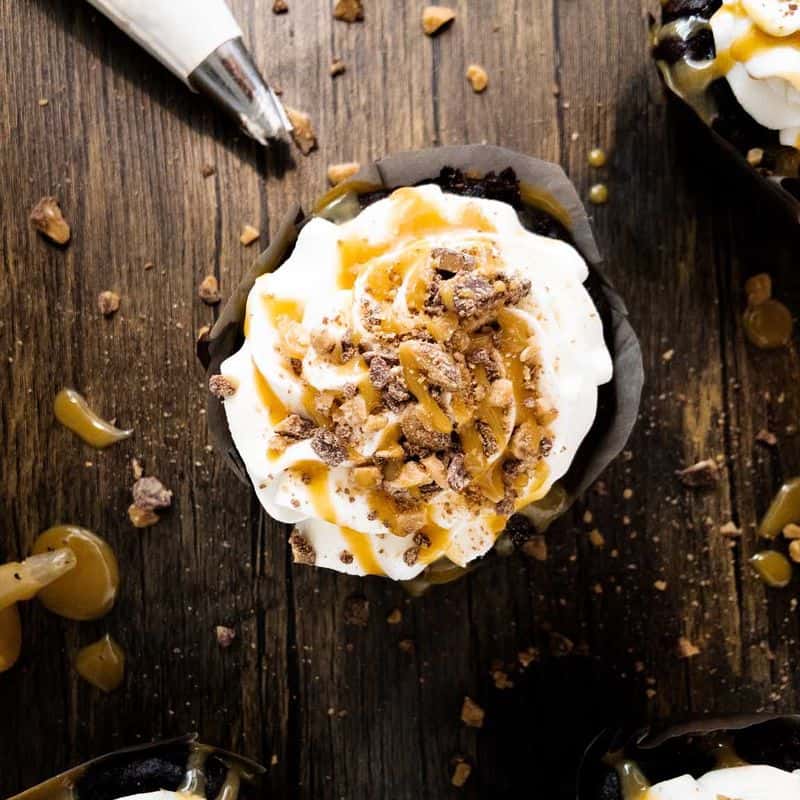 better than sex cupcakes topped with whipped cream, caramel sauce, and Heath bits, on the counter next to a piping bag filled with whipped cream