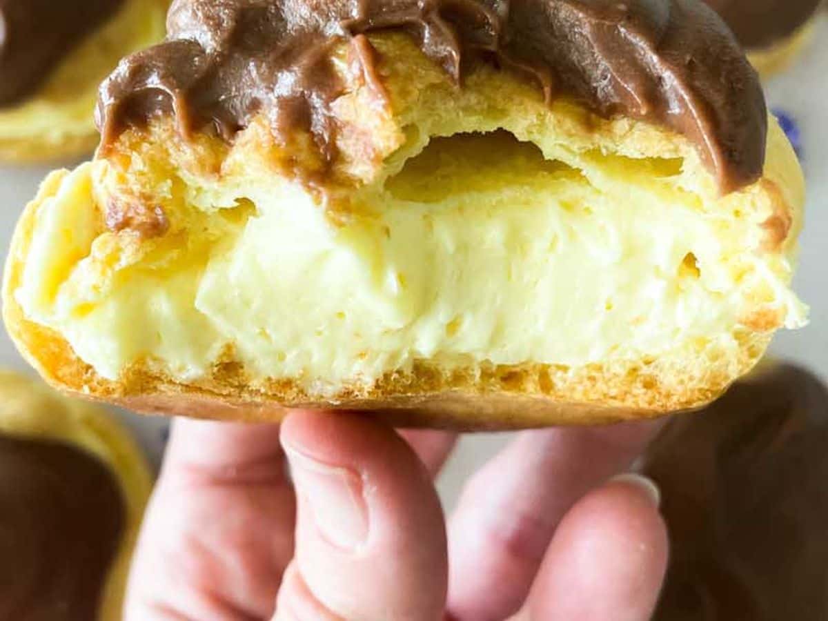 hand holding chocolate eclair with bite taken out of it and cream filling showing