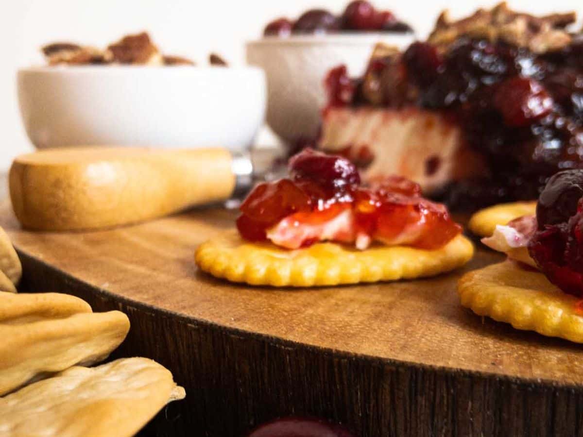 cranberry cream cheese dip on a cracker with block of dip, bowl of cranberries, and bowl of pecans in the background