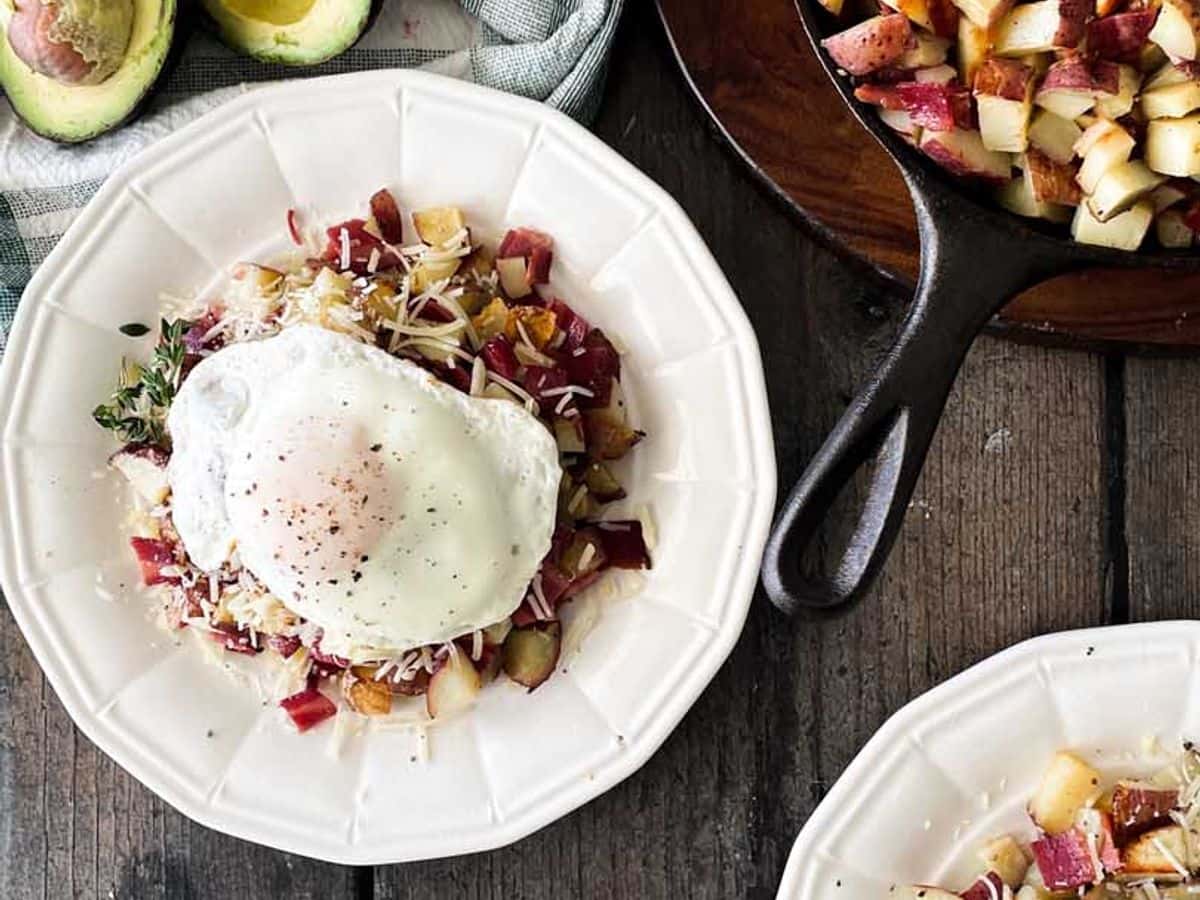 two plates of roasted potatoes and bacon hash topped with parmesan cheese and fried egg surrounded by ingredients