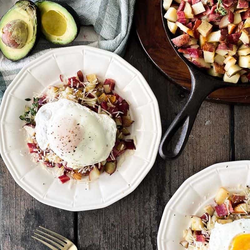two plates of roasted potatoes and bacon hash topped with parmesan cheese and fried egg surrounded by ingredients